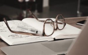 Image of a pair of brown glasses placed on a book which has lots of writing on in black ink. White pen lying on top of glasses, silver phone in the background and a silver laptop