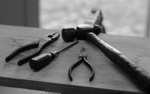 Image in black and white, of tools including a hammer, screwdriver and pliers. They are sat on a wooden plank