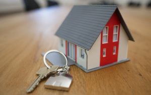 Image of a small toy house and a key with a key ring in the shape of a house, both placed on a light brown wooden table