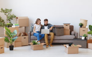 Couple who have just moved house, with moving in boxes around them. Sat on a sofa with a laptop